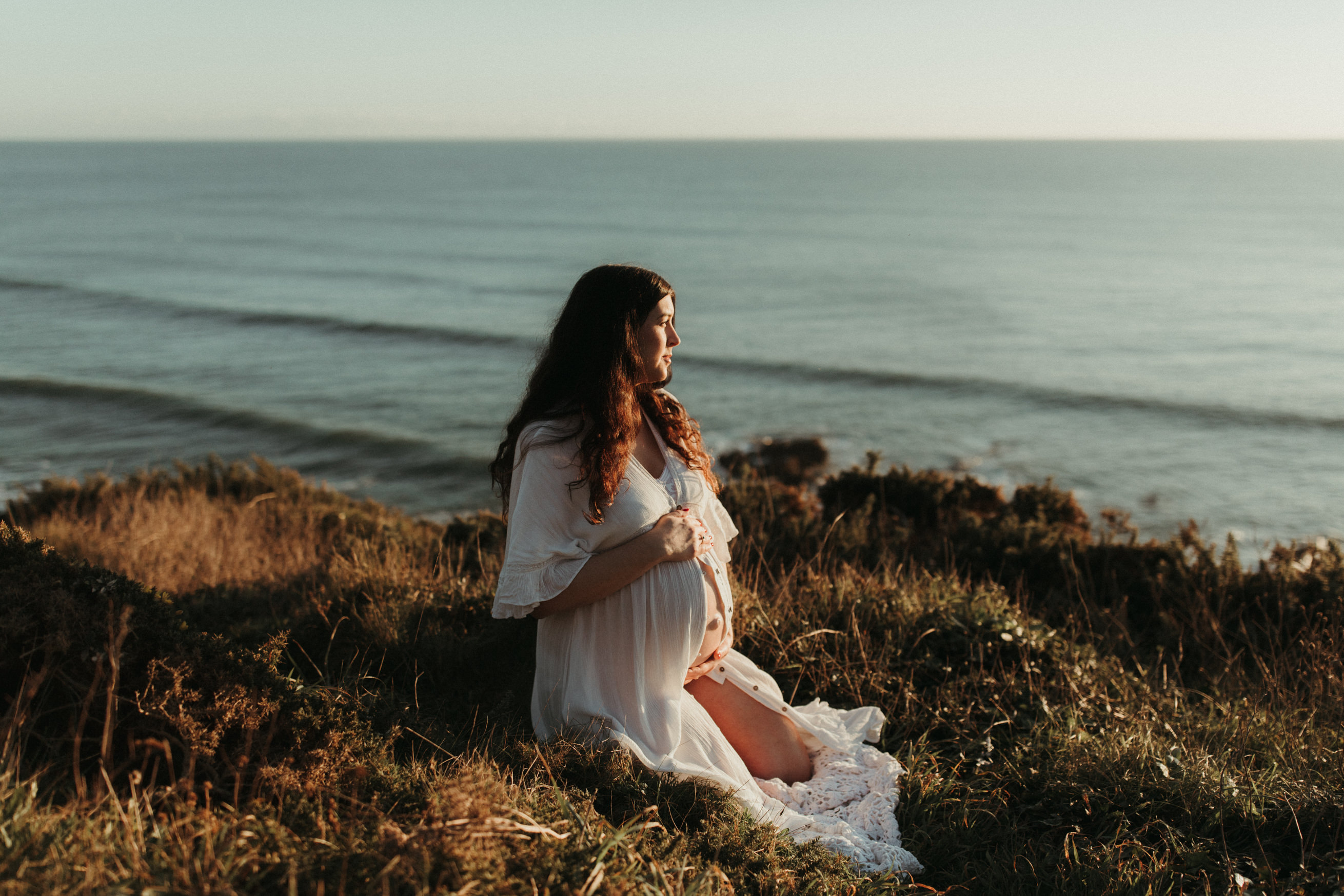 Maternity and Newborn Photographer Devon woman kneels in sand dunes and looks out to sea