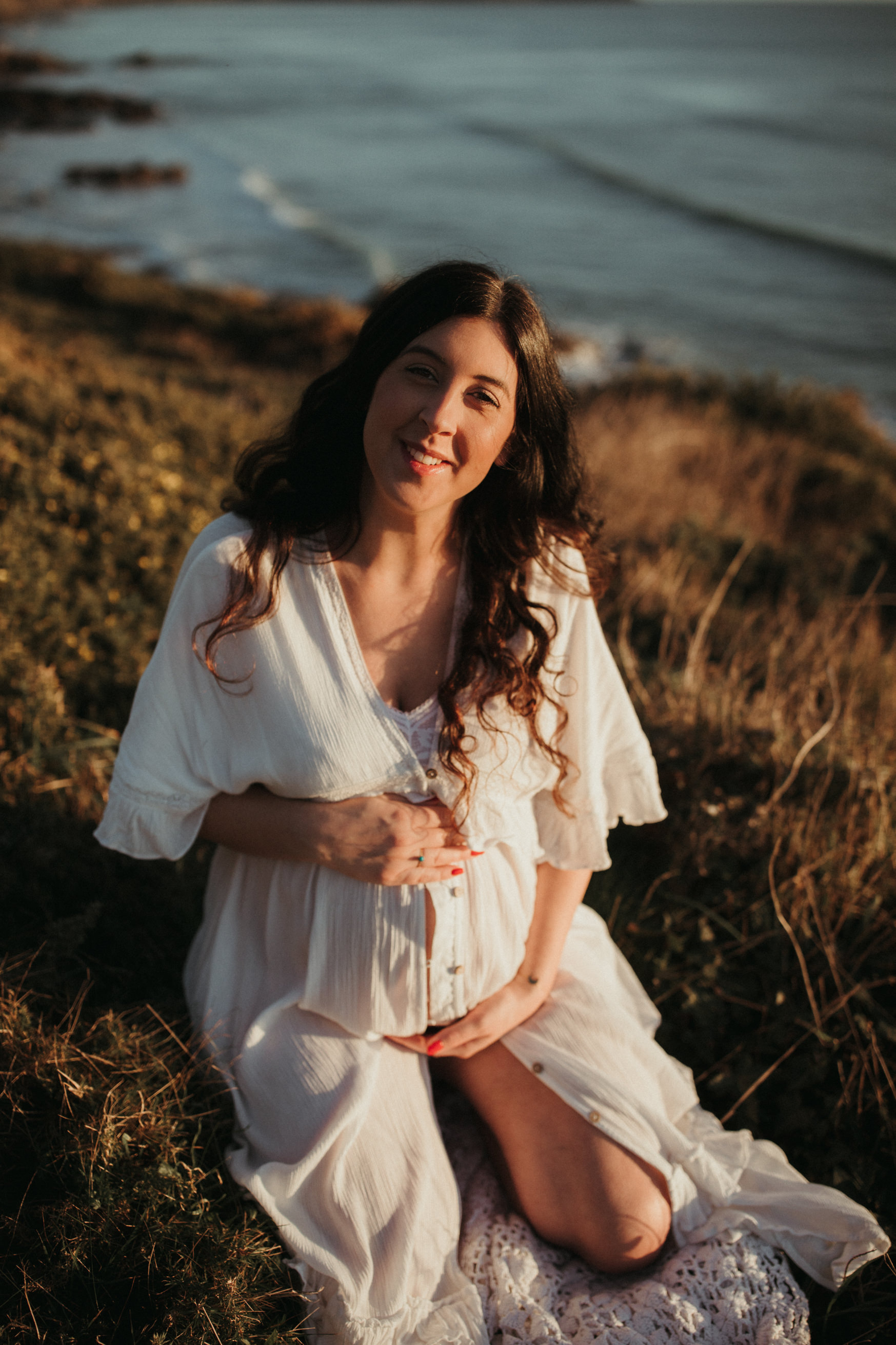 Pregnant Woman sits in sand dunes and smiles at camera Maternity and Newborn Photographer Devon 