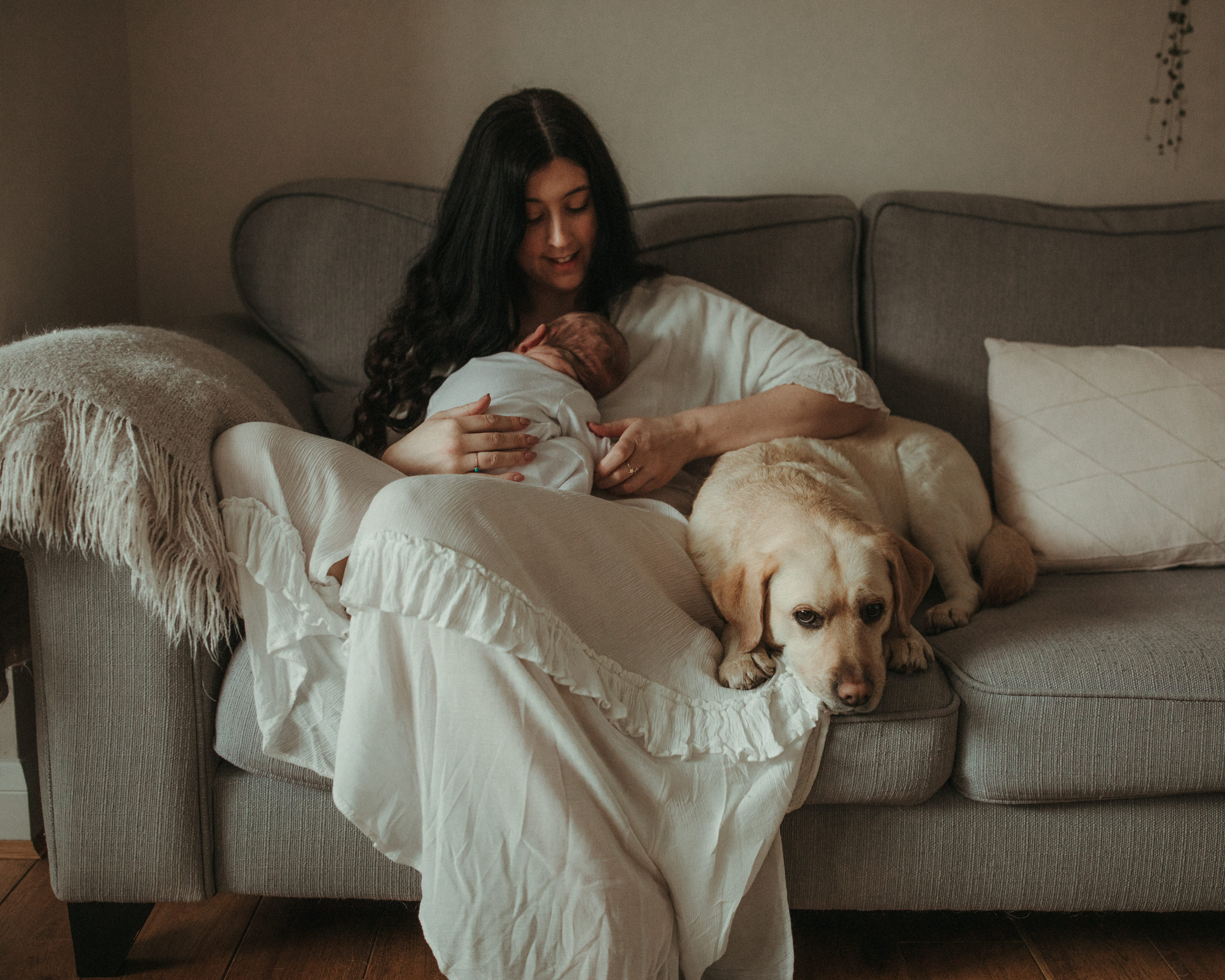 Mother sits with newborn whilst dog lies next to them on sofa Maternity and Newborn Photographer Devon 