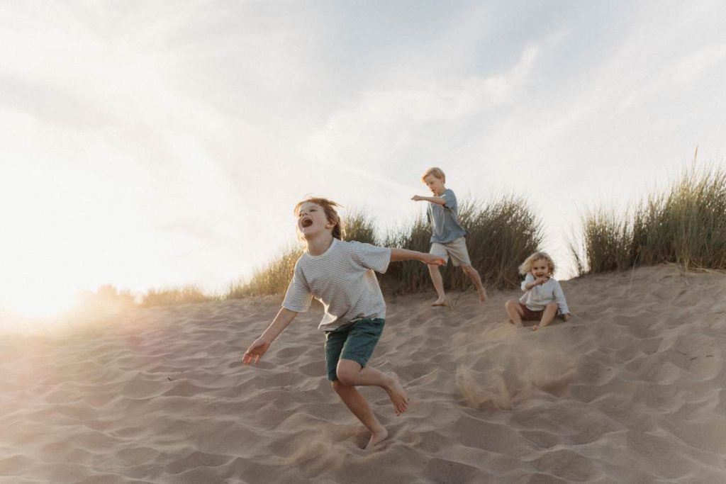 Three boys running down a sand dunes on the beach in Cornwall in the summer,. The sun is setting behind them.