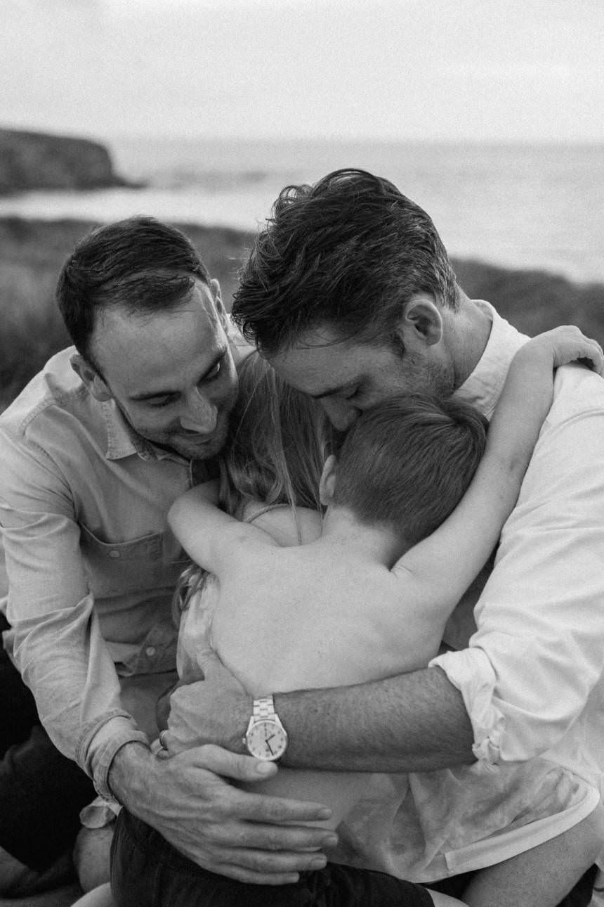 A black and white image of two fathers are embracing their children and each other, a family hug, on the beach in Cornwall.