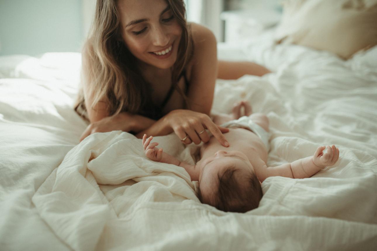 A mother lying on her bed looking down and smiling at her baby while she tickles her tummy.
