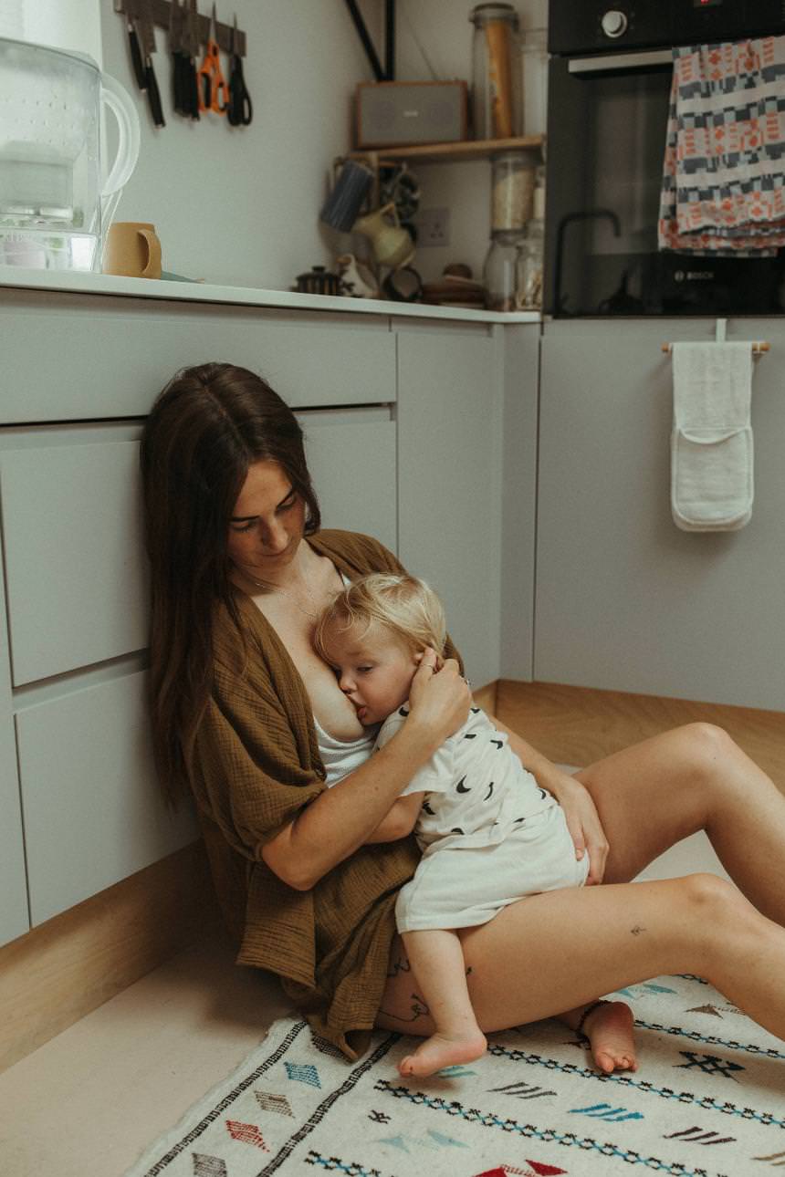 A mother is sitting down in the kitchen of her home and breastfeeding her young son.