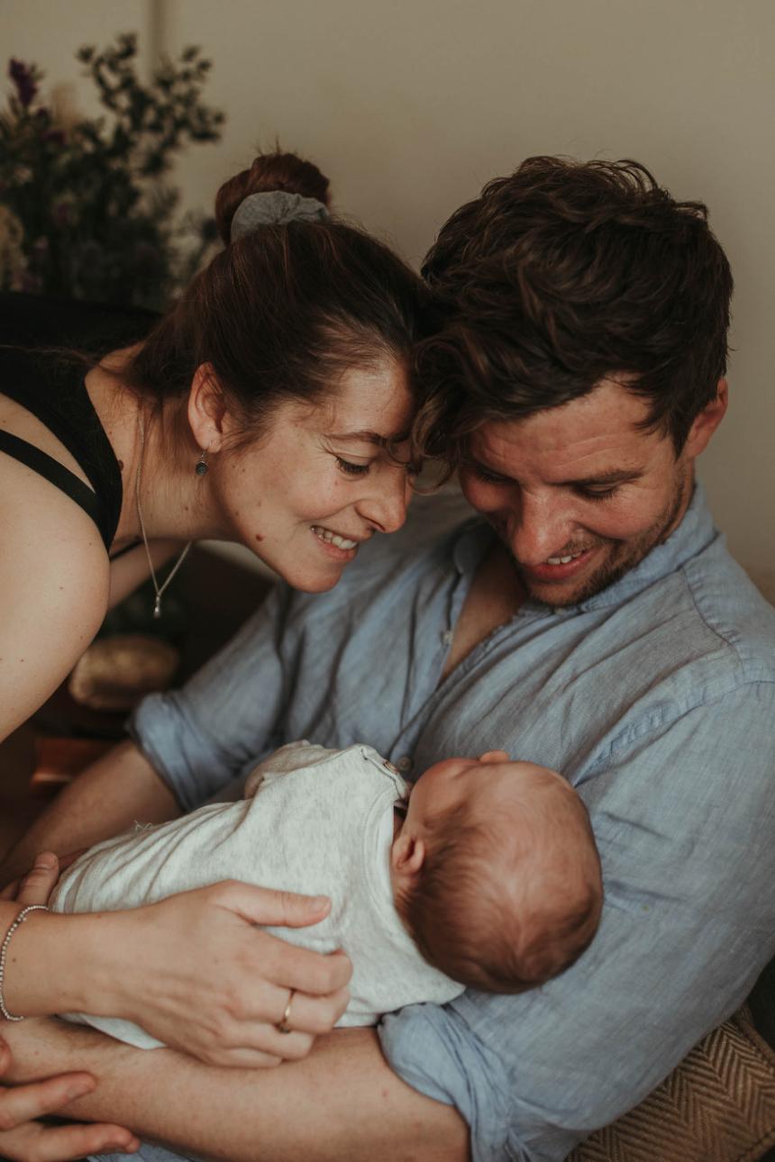 A family photo of a mother and father looking down and smiling at their newborn baby boy in their own home in Cornwall.