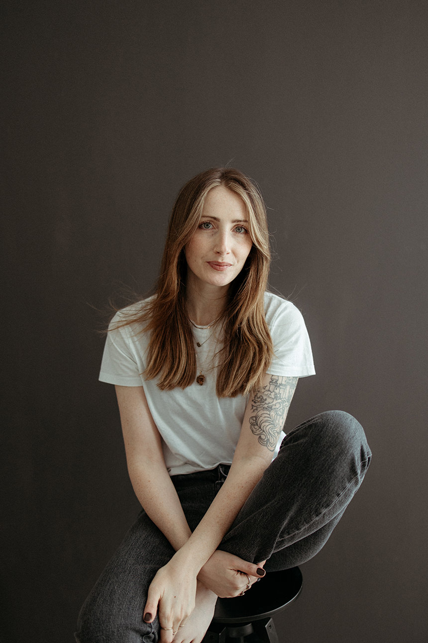 An image of the photographer behind Melanie Grace Cornwall family Photographer, sat casually on a stool infant of a dark grey wall.
